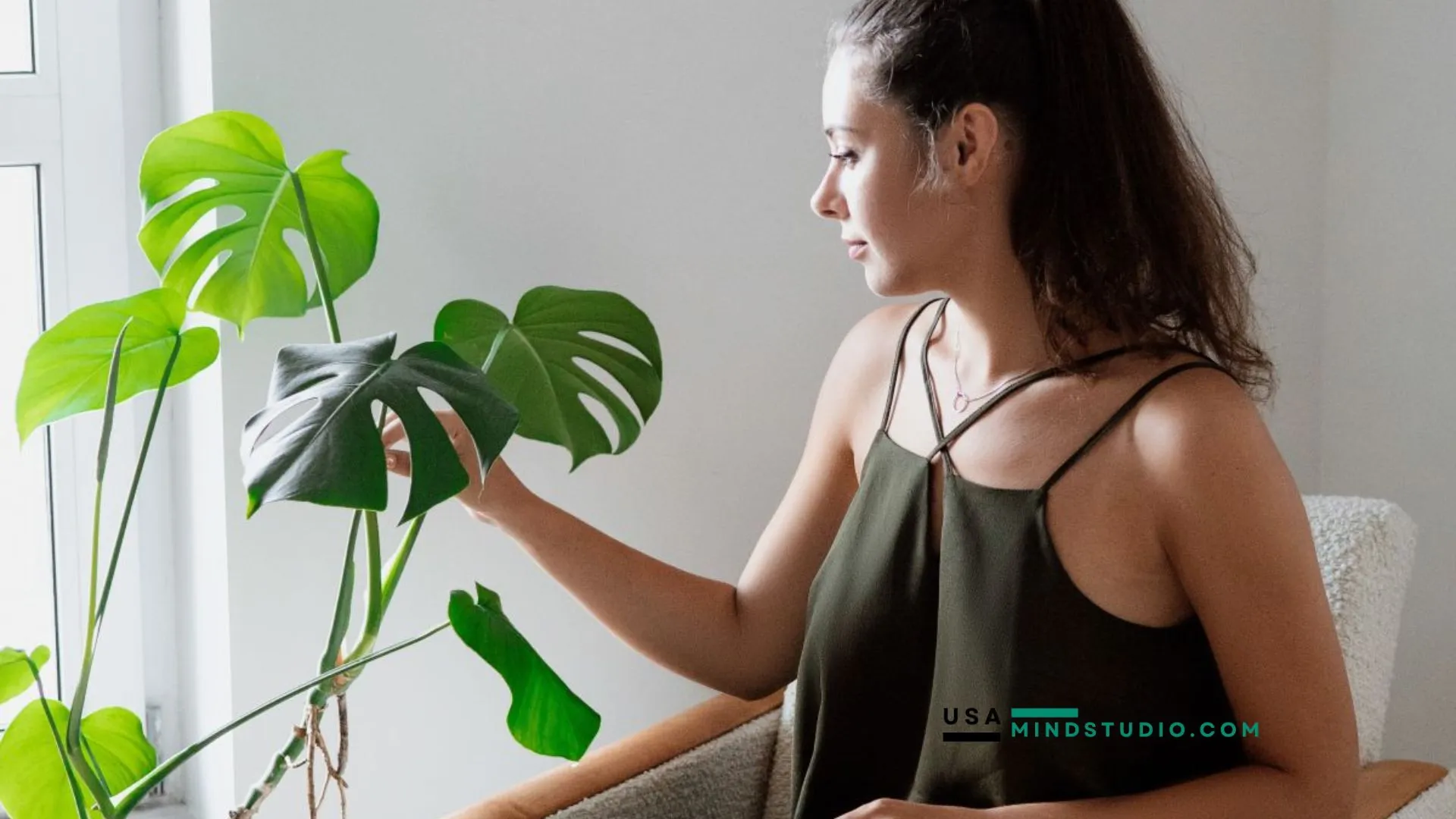 A woman sits by a window, surrounded by natural light, with a green plant beside her, creating a serene atmosphere.